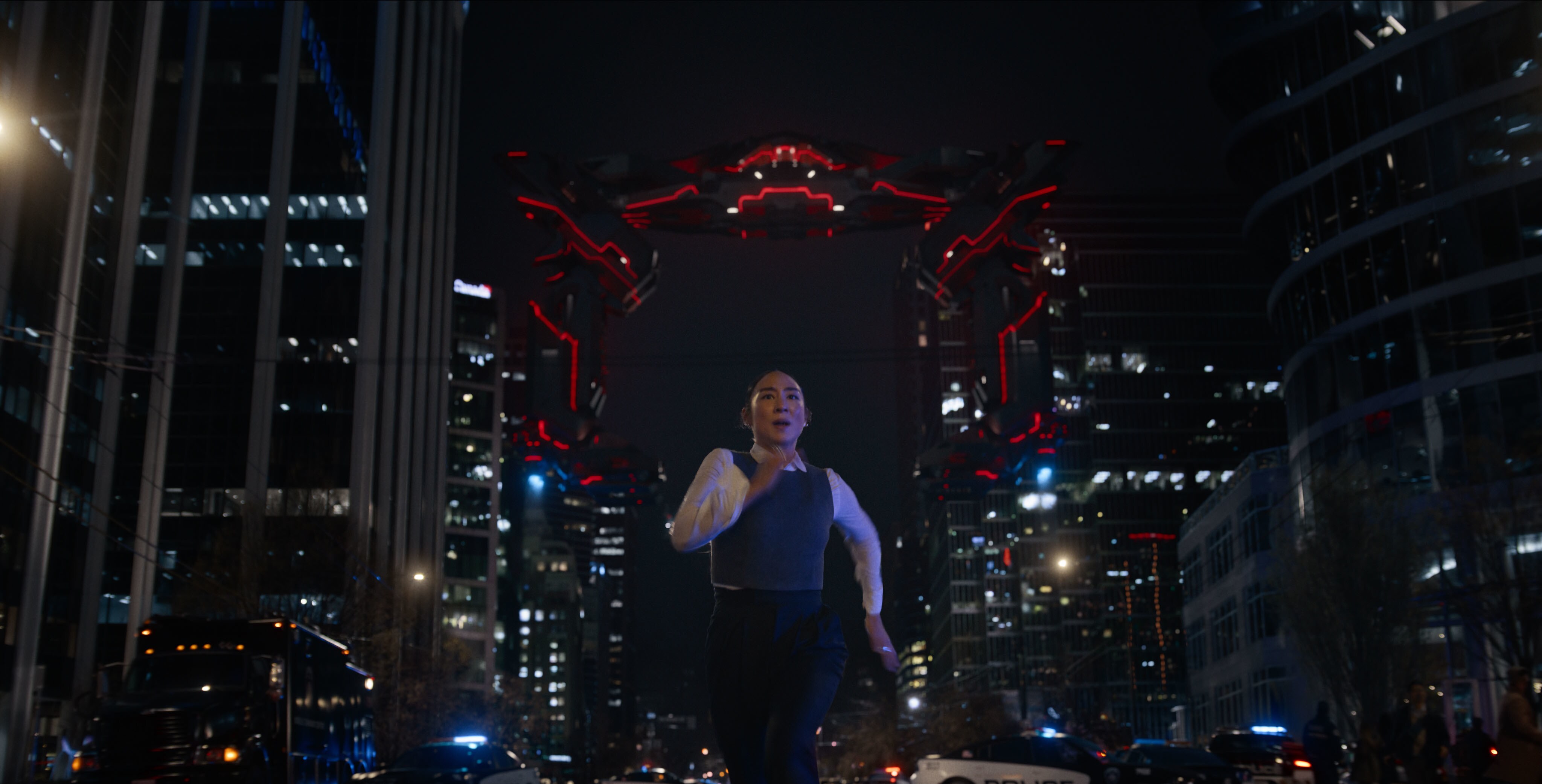 An Asian-American woman dressed in business casual runs down a street towards the camera, chased by a giant red-and-black geometrical spaceship. The overall shot looks surprisingly cheap.