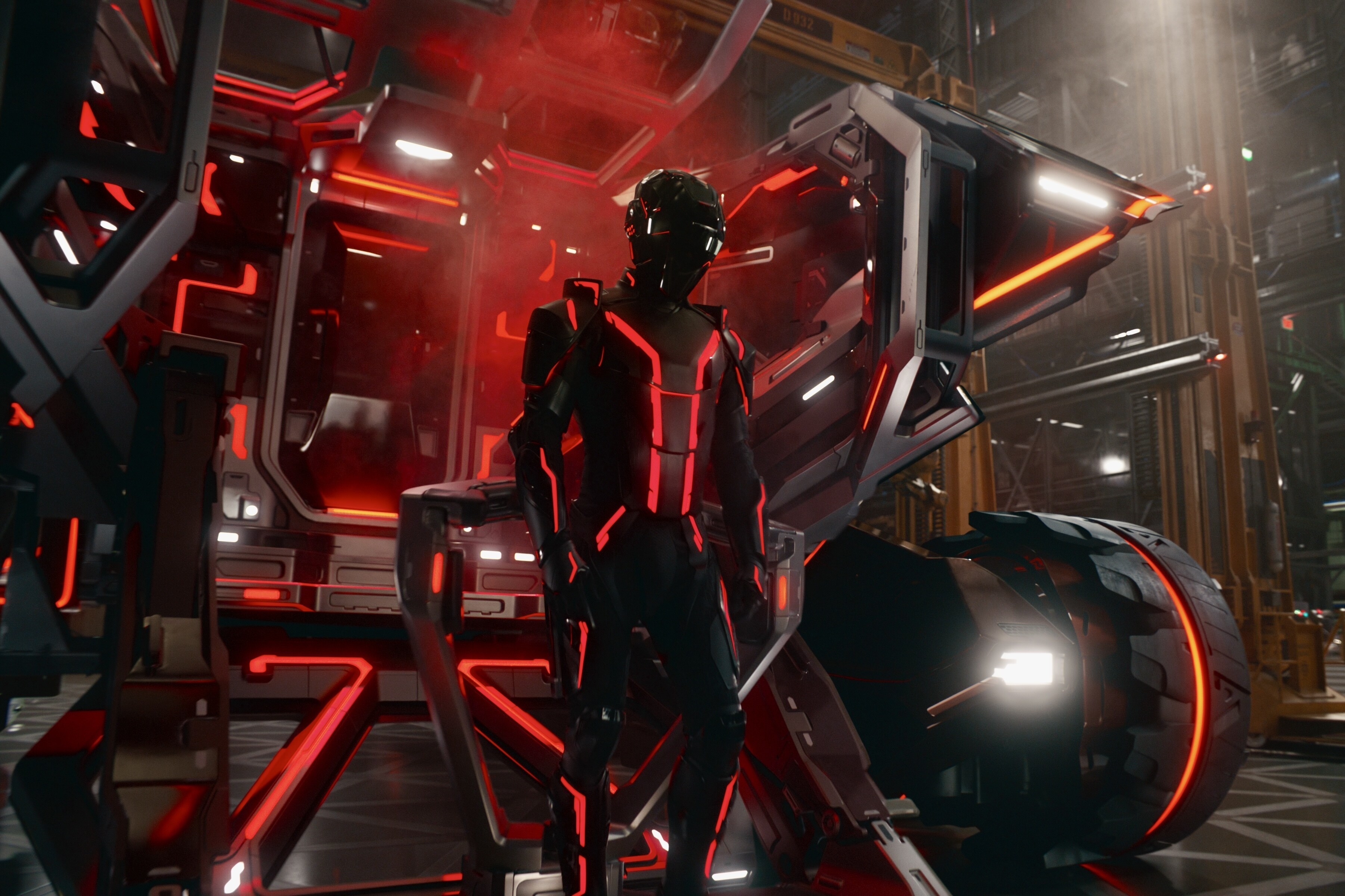 On a factory floor, a human figure clad in black spandex armor with neon red lights and a glossy blackout helmet stands in front of a very sci-fi vehicle with large tires and an open back. The shot feels oddly cheap despite its technical sophistication.