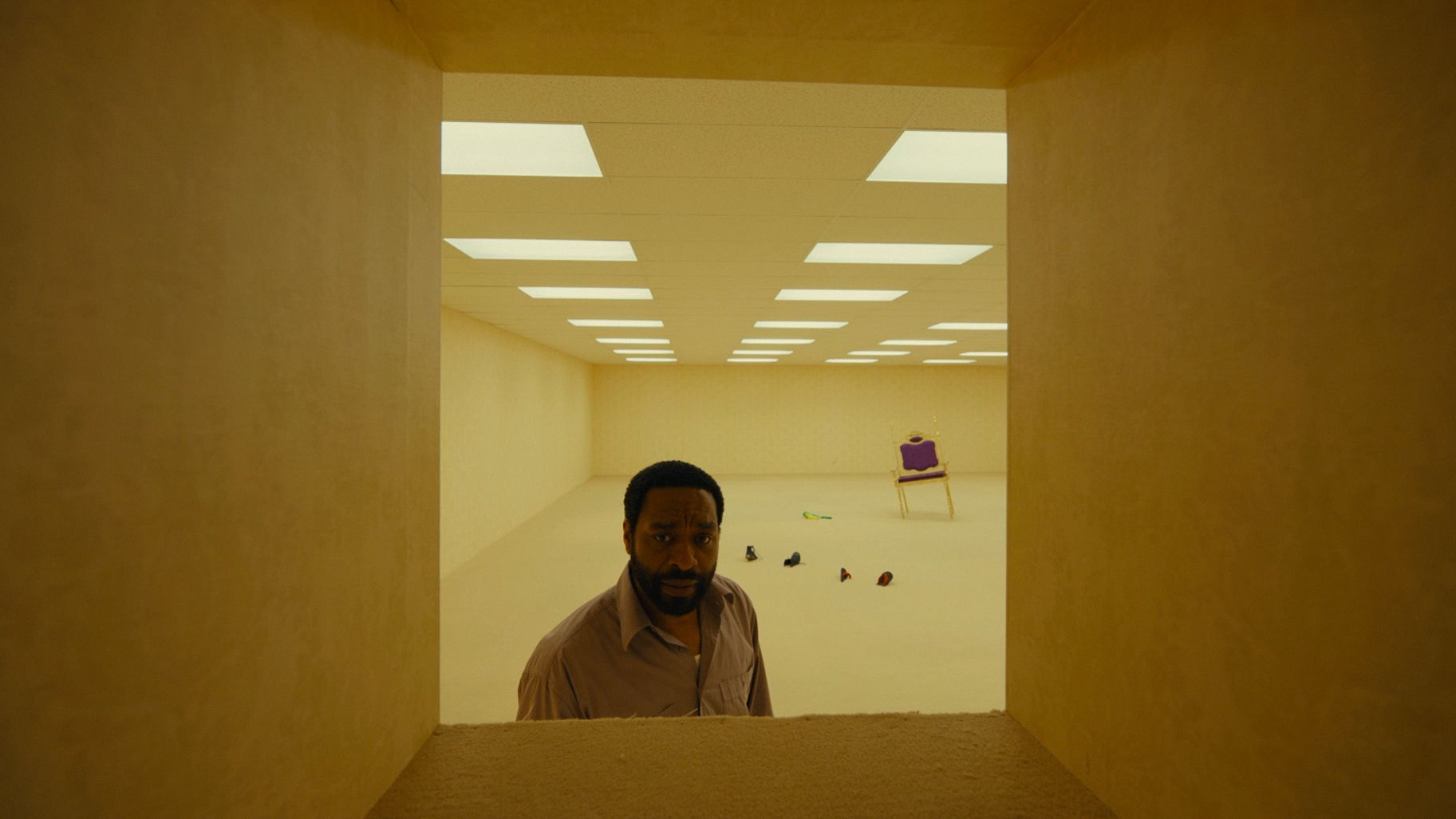 An all-light-yellow room with wallpaper, carpeted floors and square light tiles regularly spaced on a drop ceiling. We are looking at it through an also-wallpapered also-carpeted square tunnel barely wide enough for an adult human to pass through. A Black man looks back through the tunnel. Behind him is a tacky throne-like chair, half-sunken into the carpet, along with two pairs of shoes.